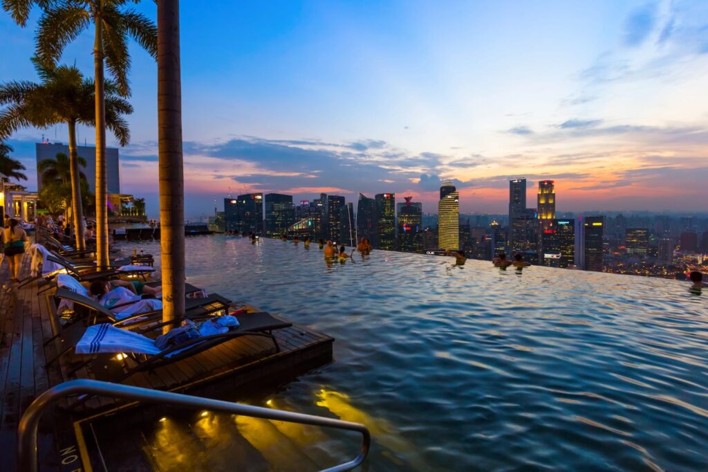 The evening city view features rows of skyscrapers, a swimming pool, and palm trees.